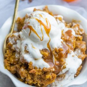 One serving of a pumpkin cobbler topped with vanilla ice cream, caramel sauce in a white bowl with a gold spoon