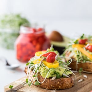 Vertical view of an open-faced rainbow vegetable sandwich topped with avocado and fresh vegetables on a wooden cutting board