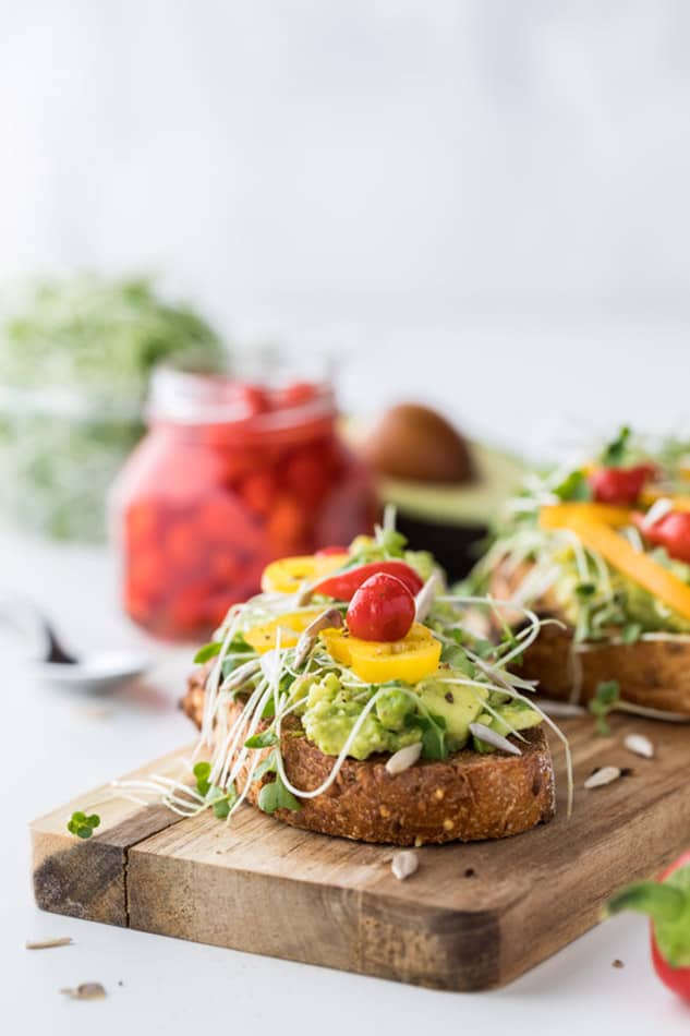 Vertical view of an open-faced rainbow vegetable sandwich topped with avocado and fresh vegetables on a wooden cutting board
