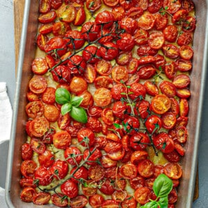 A batch of caramelized roasted cherry tomatoes on a baking sheet with basil and fresh herbs