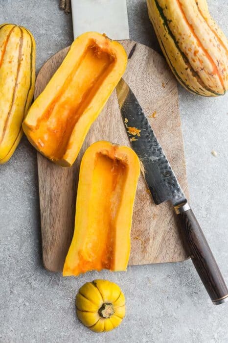 Top view of raw delicata squash cut into halves lengthwise on a wooden cutting board with a knife