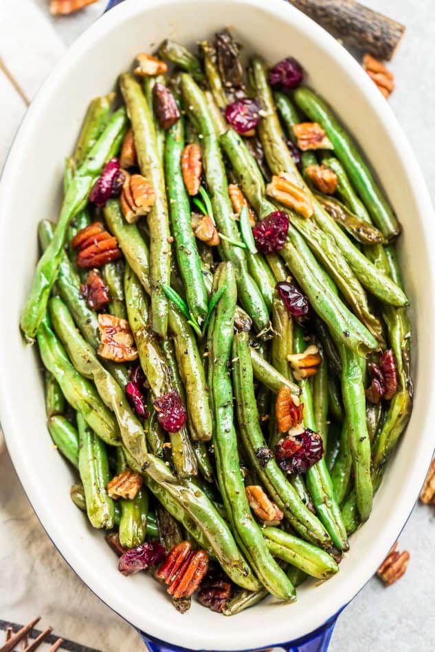 An oval serving dish with roasted green beans, topped with pecans and dried cranberries