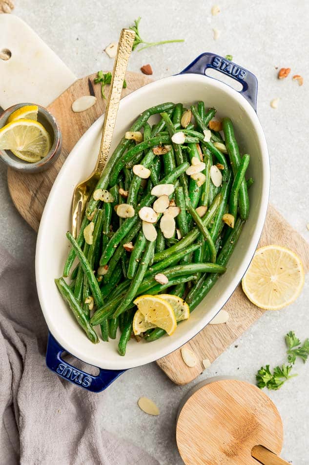 An oval dish with roasted green beans topped with lemon slices and shaved almonds, on a cutting board next to lemon slices and parsley