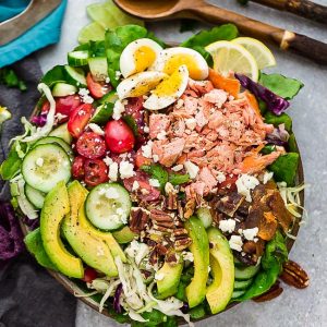 Top view of fresh salmon salad in a white bowl on a light grey background with a fork on the counter below it and a wooden serving spoon.