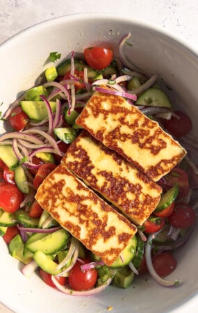 Three rectangle slabs of seared Halloumi cheese over a bed of mixed salad greens in a white bowl