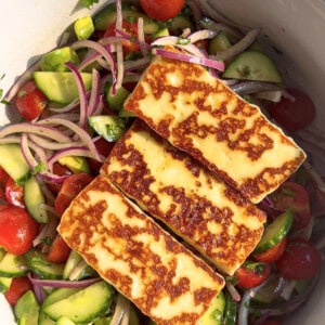 Three rectangle slabs of seared Halloumi cheese over a bed of mixed salad greens in a white bowl