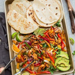 A pair of tongs grabbing a scoop of fajita fillings from a sheet pan with warm tortillas beside the meat and veggies