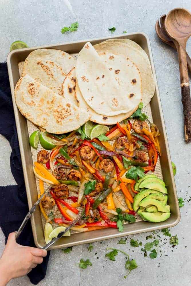 someone using tongs to remove cooked shrimp and vegetables from a Sheet Pan Shrimp Fajitas meal