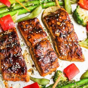 Three baked teriyaki salmon fillets on a baking sheet surrounded by asparagus and bell peppers