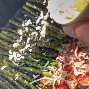 Shredded mozzarella being sprinkled on asparagus and tomatoes in an air fryer