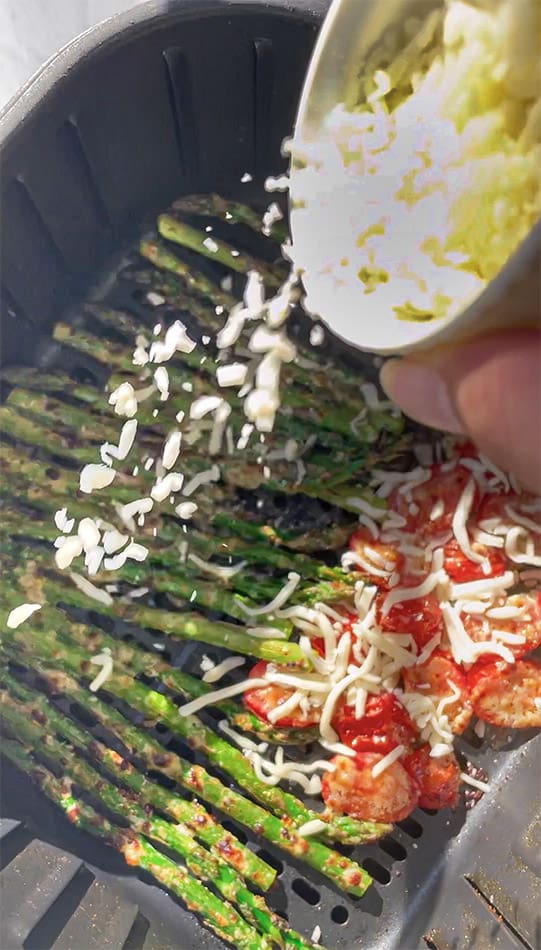 Shredded mozzarella being sprinkled over tomatoes and asparagus in an air fryer