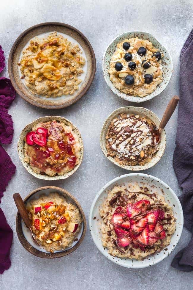 Top view of six basic oatmeal bowls with different toppings on a grey background with a spoon and napkins