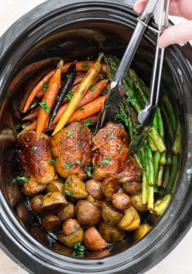 An entire batch of Slow Cooker Chicken Dinner: four chicken thighs, baby potatoes, asparagus and baby carrots in a black slow cooker with tongs.