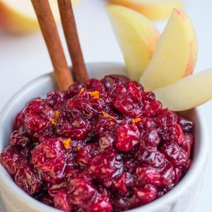 Close-up of a bowl of homemade cranberry sauce with 2 cinnamon sticks and apple slices