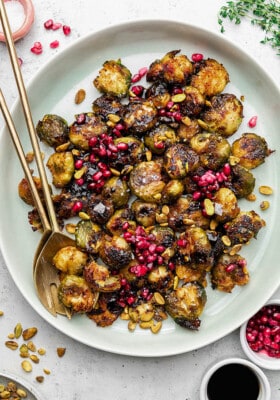 Close-up top shot of a white bowl filled with roasted and smashed brussels sprouts topped with pomegranate seeds and pistachios with two golden spoons