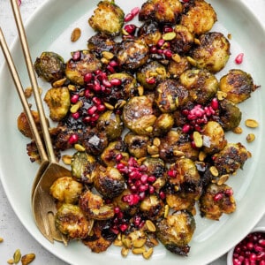 Close-up top shot of a white bowl filled with roasted and smashed brussels sprouts topped with pomegranate seeds and pistachios with two golden spoons