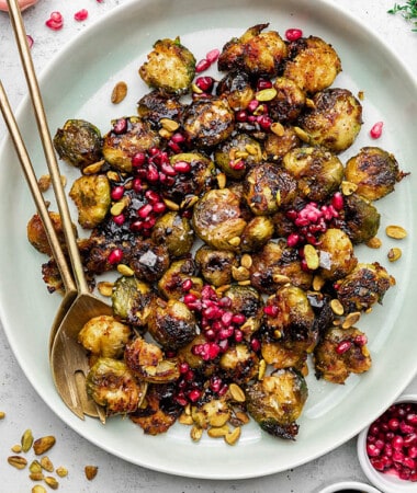 Close-up top shot of a white bowl filled with roasted and smashed brussels sprouts topped with pomegranate seeds and pistachios with two golden spoons