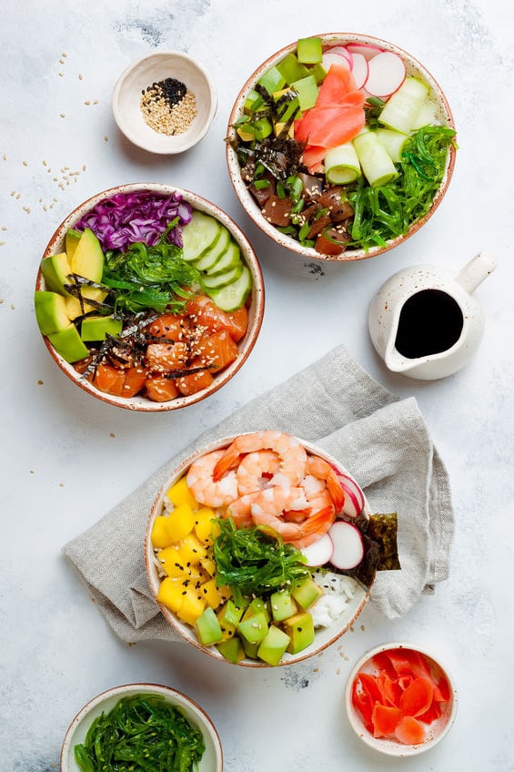 Hawaiian salmon, tuna and shrimp bowls on a countertop with seaweed, avocado, mango, pickled ginger and sesame seeds