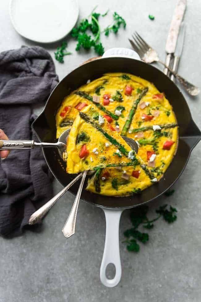 Top view of Keto Frittata with Asparagus, Tomatoes and Ham in a White Cast Iron Skillet Pan with a missing slice held up with two forks