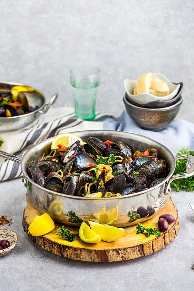 Steamed Mediterranean PEI Mussels in a skillet with lemon wedges and olives on a piece of wood