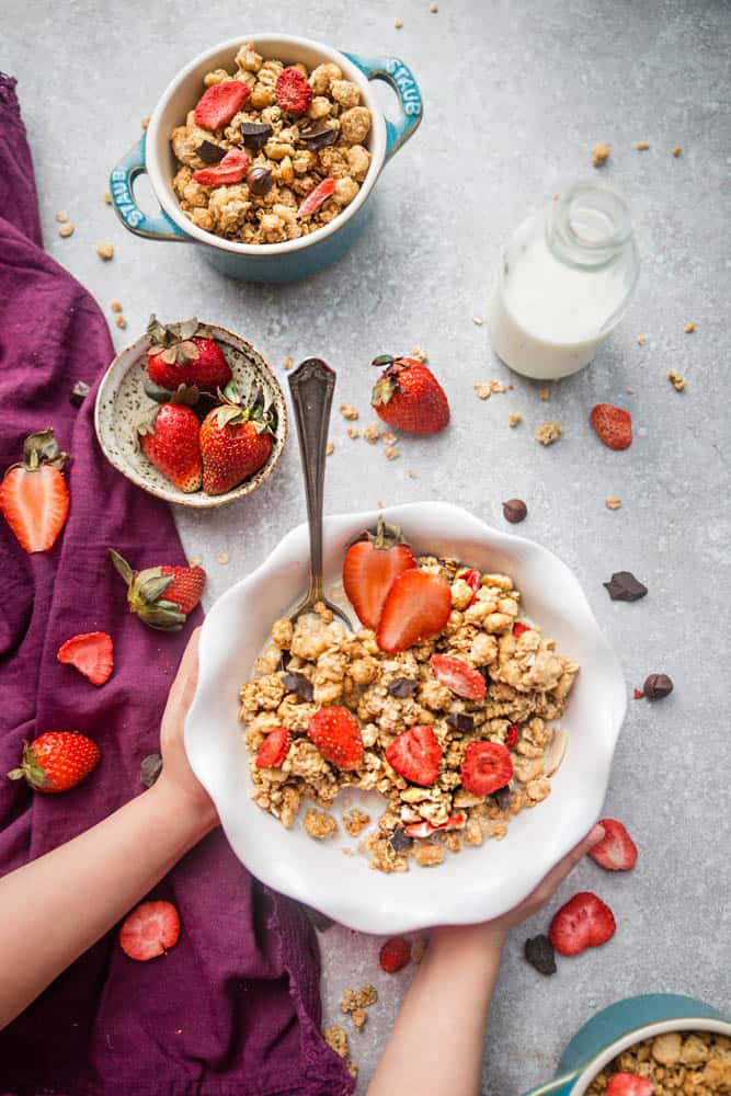 Two hands holding a bowl of strawberry chocolate granola with milk, topped with fresh strawberries, next to a table full of strawberries, a bowl of strawberries, another bowl of granola, and a bottle of milk