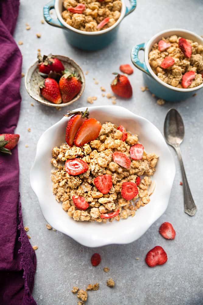 A bowl of strawberry granola in a white bowl with a spoon on a grey background