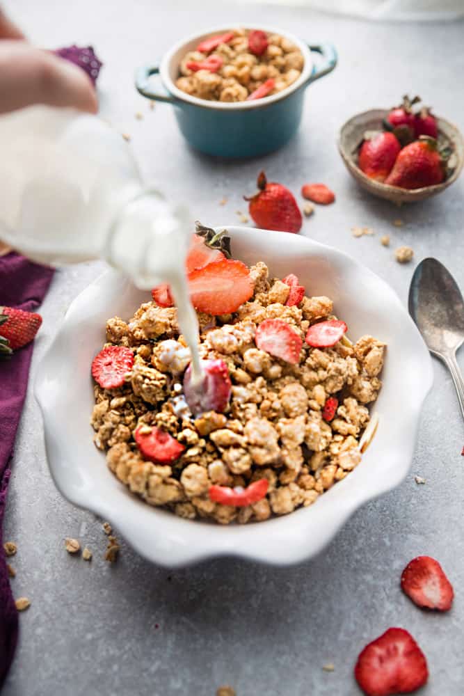 A bowl of strawberry granola in a white bowl with fresh strawberries on a grey background with milk pouring into the bowl
