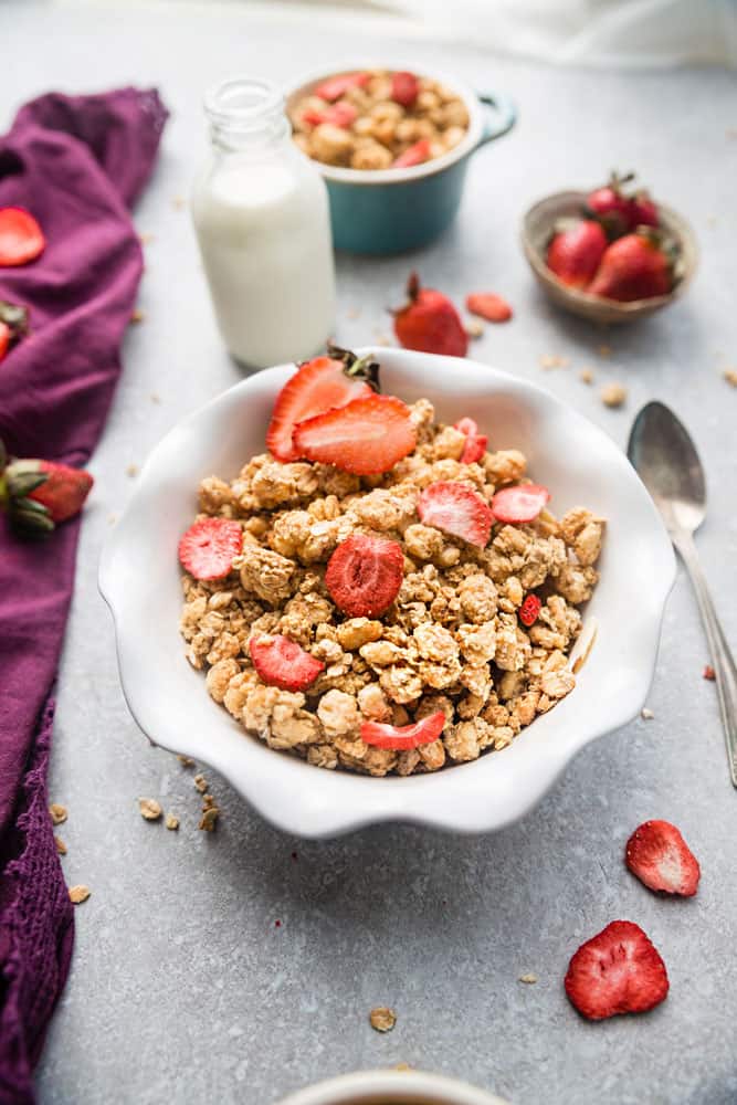 A bowl of strawberry granola in a white bowl with a spoon on a grey background