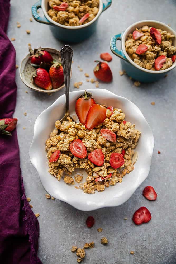 A bowl of strawberry chocolate granola with milk in it, topped with fresh strawberries, with a spoon in the bowl, with more bowls of granola in the background, and a bull of whole strawberries