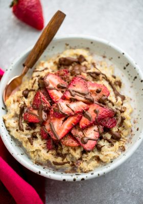 Top view of strawberry oatmeal in a white speckled bowl with a spoon on a grey background and a red napkin