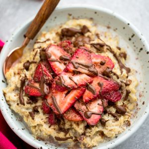 Top view of strawberry oatmeal in a white speckled bowl with a spoon on a grey background and a red napkin