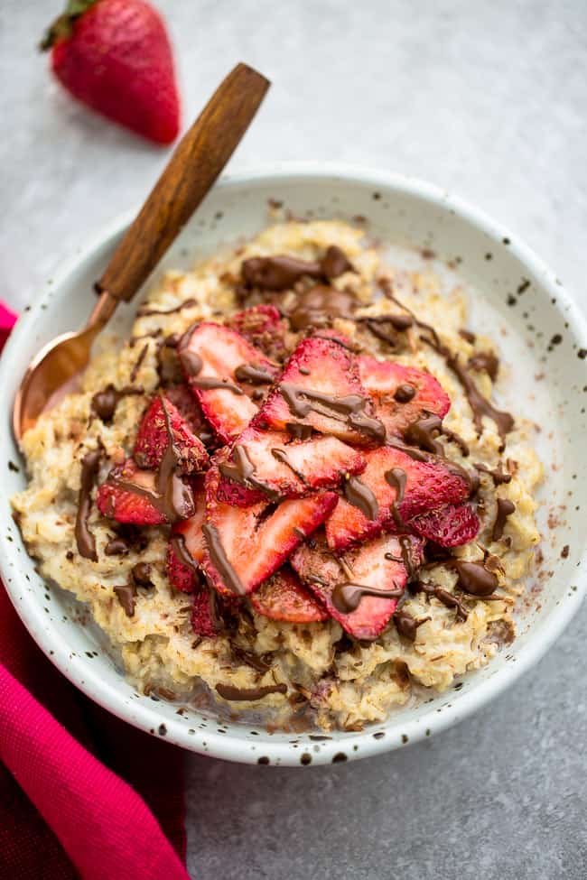 Top view of strawberry oatmeal in a white speckled bowl with a spoon on a grey background and a red napkin