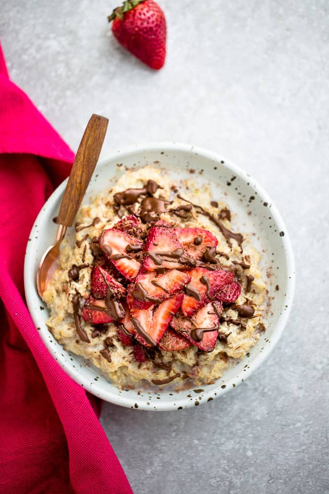 Top view of strawberry oatmeal in a white speckled bowl with a spoon on a grey background and a red napkin