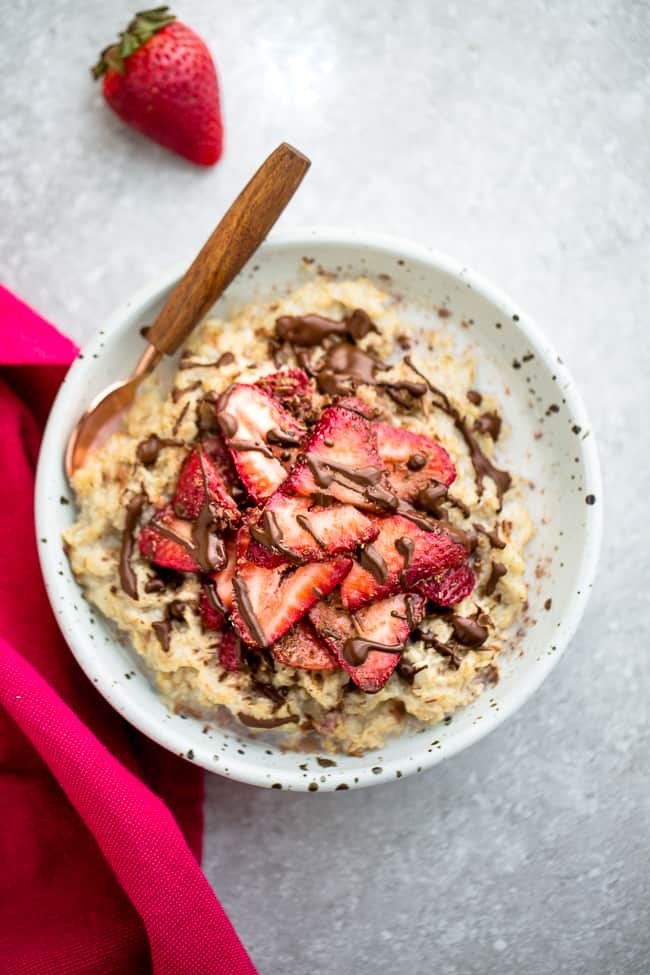 Top view of strawberry oatmeal in a white speckled bowl with a spoon on a grey background and a red napkin