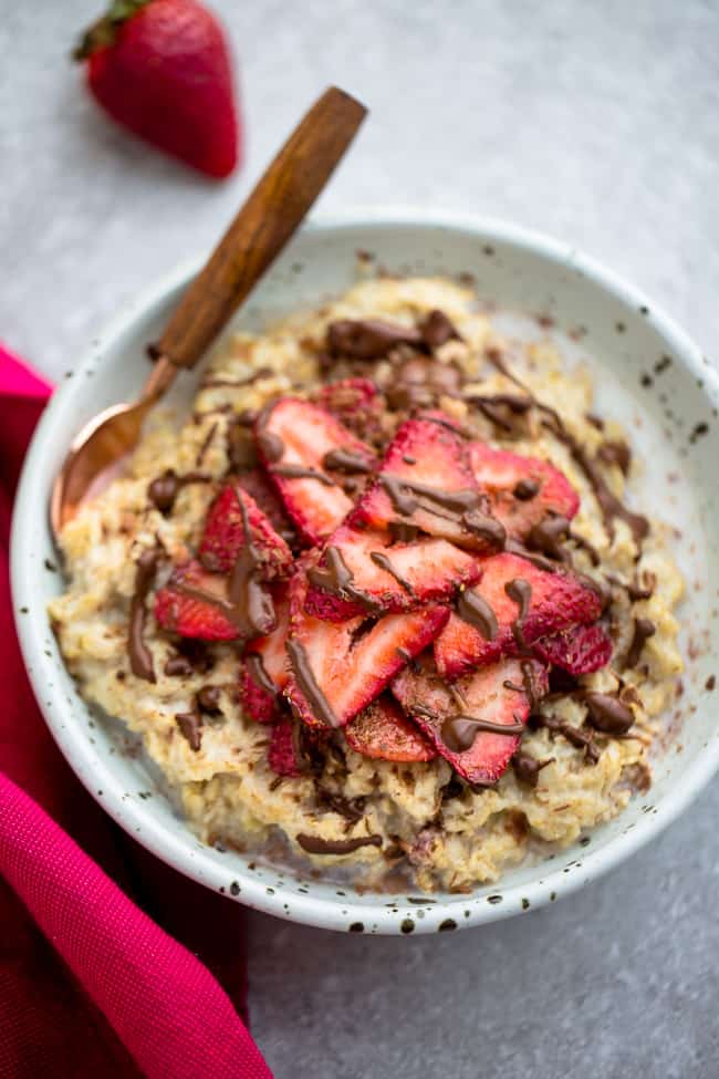 Top view of strawberry oatmeal in a white speckled bowl with a spoon on a grey background and a red napkin