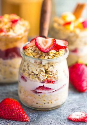 Side view strawberry overnight oats with strawberries with a spoon on a grey background with other overnight oats flavors in the background