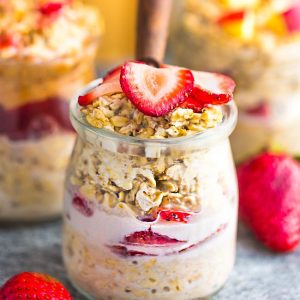 Side view strawberry overnight oats with strawberries with a spoon on a grey background with other overnight oats flavors in the background