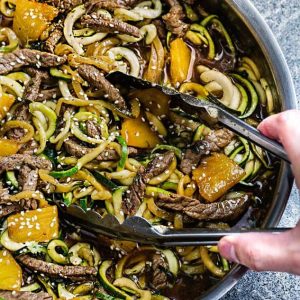 Metal tongs reaching into a skillet of Teriyaki Beef Stir-Fry with Zucchini Noodles