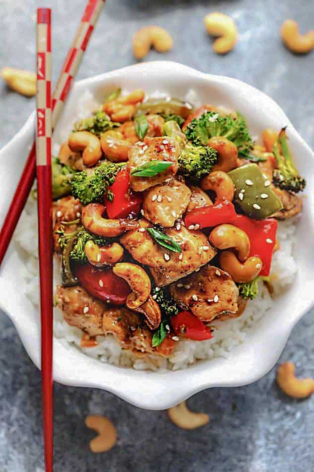 A bowl of white rice with stir-fried chicken and veggies on top and a pair of chopsticks resting on the rim of the bowl