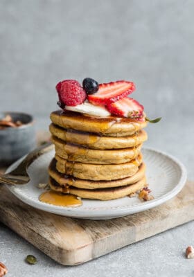 Side view of stack of fluffy almond flour pancakes with strawberries, blueberries, raspberries and sugar free maple syrup on a white plate with a gold fork