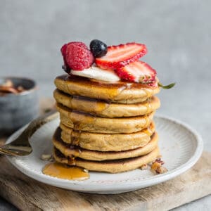 Side view of stack of fluffy almond flour pancakes with strawberries, blueberries, raspberries and sugar free maple syrup on a white plate with a gold fork