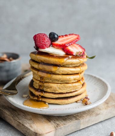 Side view of stack of fluffy almond flour pancakes with strawberries, blueberries, raspberries and sugar free maple syrup on a white plate with a gold fork