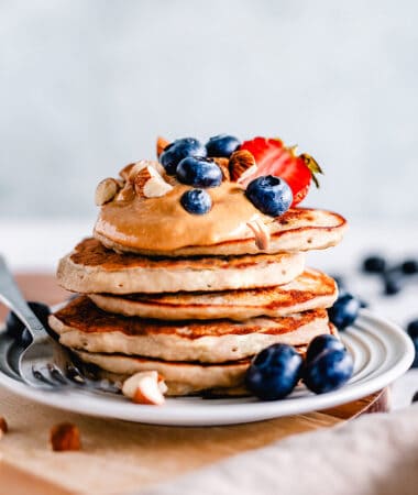 Side view of a stack of fluffy keto pancakes on a white plate with a fork and berries