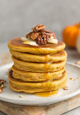 Side view of a stack of 5 thick and fluffy keto pancakes on a white plate with sugar free syrup, pecans and butter