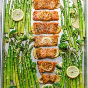 Roasted salmon fillets on a baking sheet with lemon slices and roasted veggies on the side