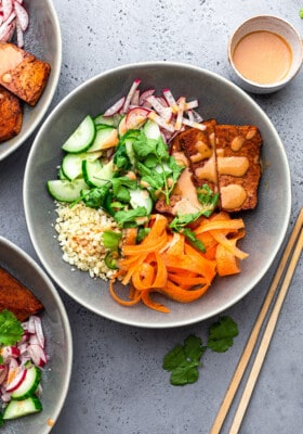 A serving of vegan bánh mi bowl topped with ribboned carrots, cauliflower rice, crispy tofu and fresh herbs in a grey bowl with chopsticks on the right side