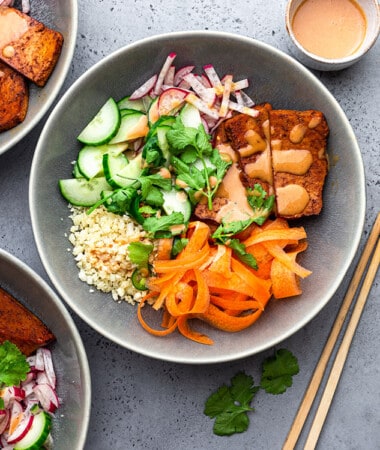 A serving of vegan bánh mi bowl topped with ribboned carrots, cauliflower rice, crispy tofu and fresh herbs in a grey bowl with chopsticks on the right side