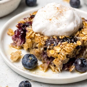 A close-up shot of a square of blueberry baked oatmeal on a white plate with a dollop of dairy-free yogurt on top