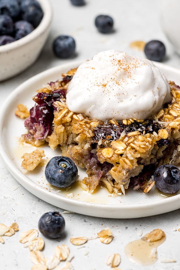 A close-up shot of a square of blueberry baked oatmeal on a white plate with a dollop of dairy-free yogurt on top