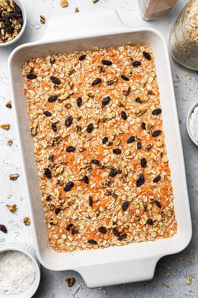 A casserole pan full of carrot cake oatmeal batter topped with chopped nuts and raisins